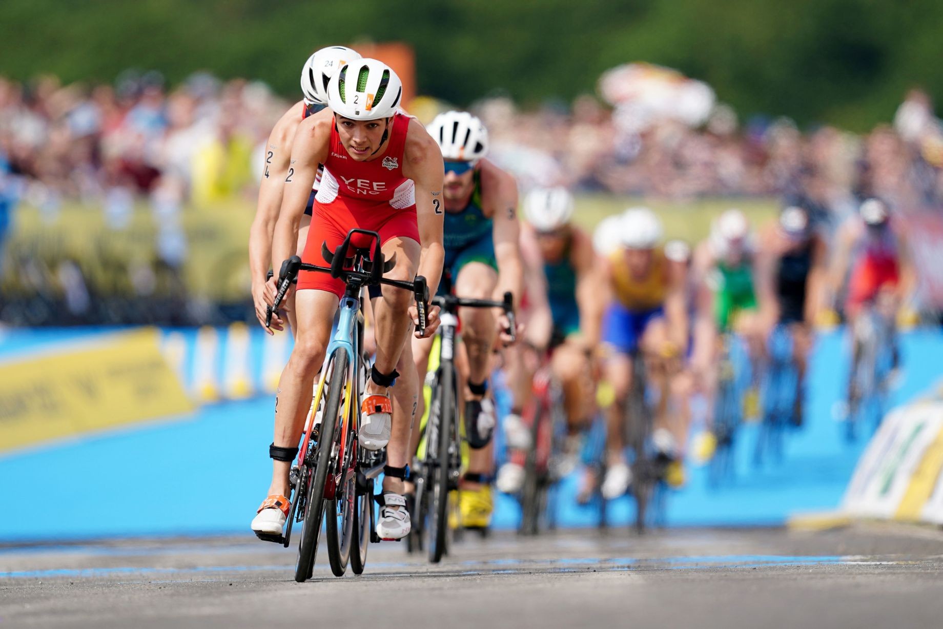 Cyclists race on an outdoor velodrome track, leaning forward on bicycles during competition.