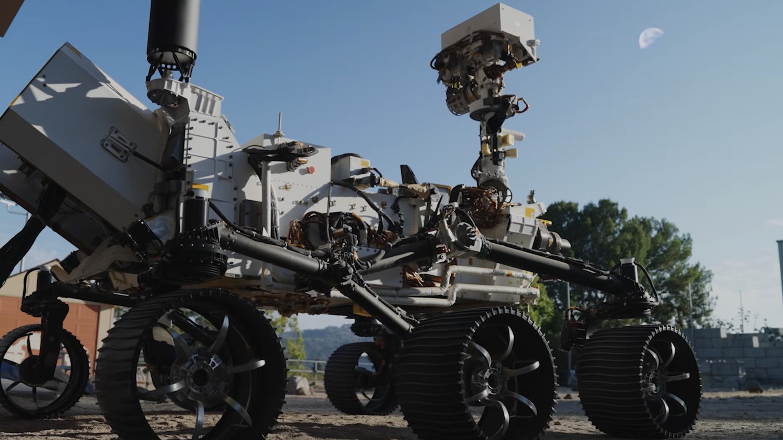 A Mars rover sits stationary on dusty ground beneath a clear blue sky with trees visible. A Mars rover sits stationary on dusty ground beneath a clear blue sky with trees visible.
