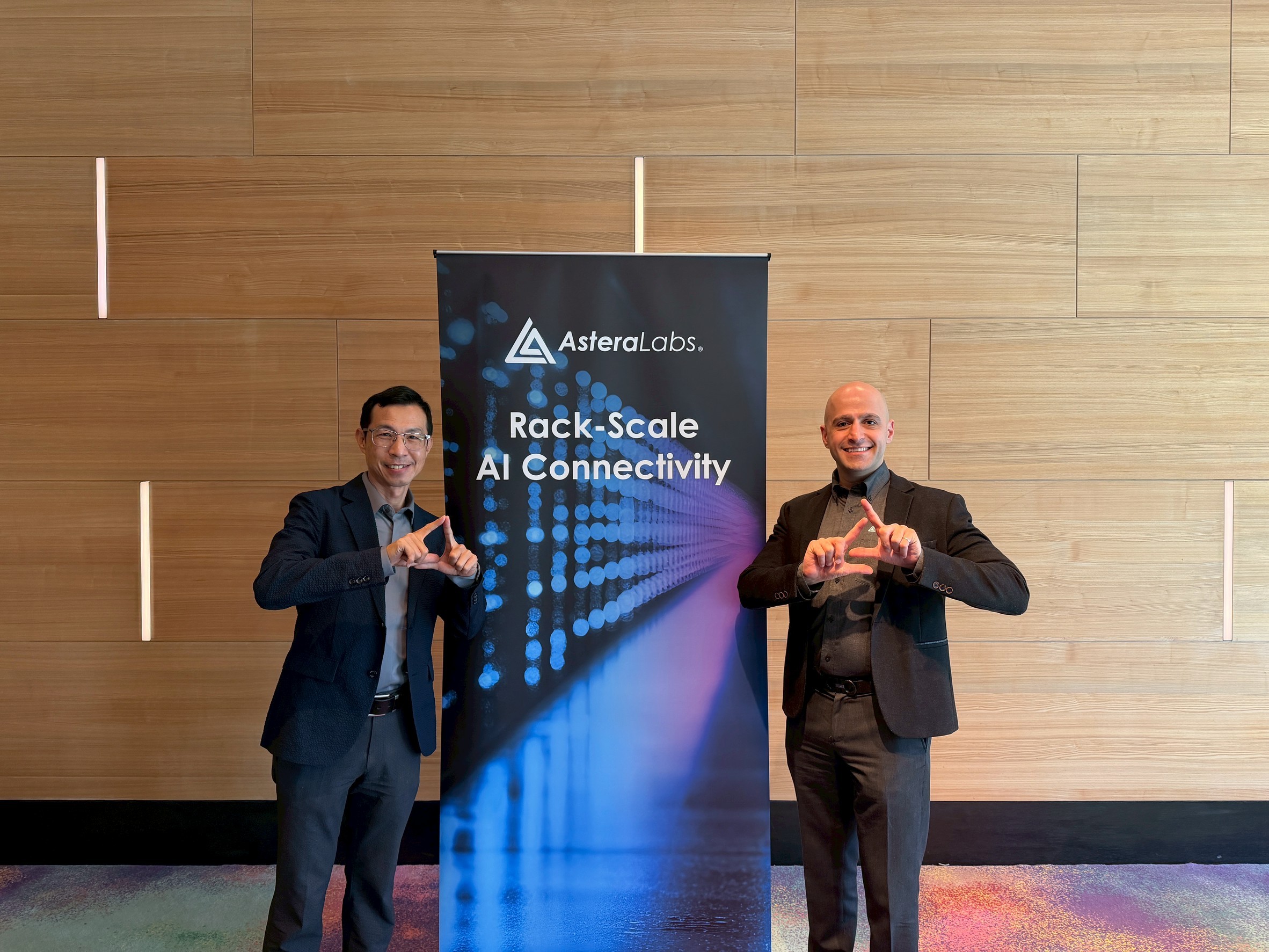 Two men pose beside an AsterLabs "Rack-Scale AI Connectivity" banner against a wooden wall.