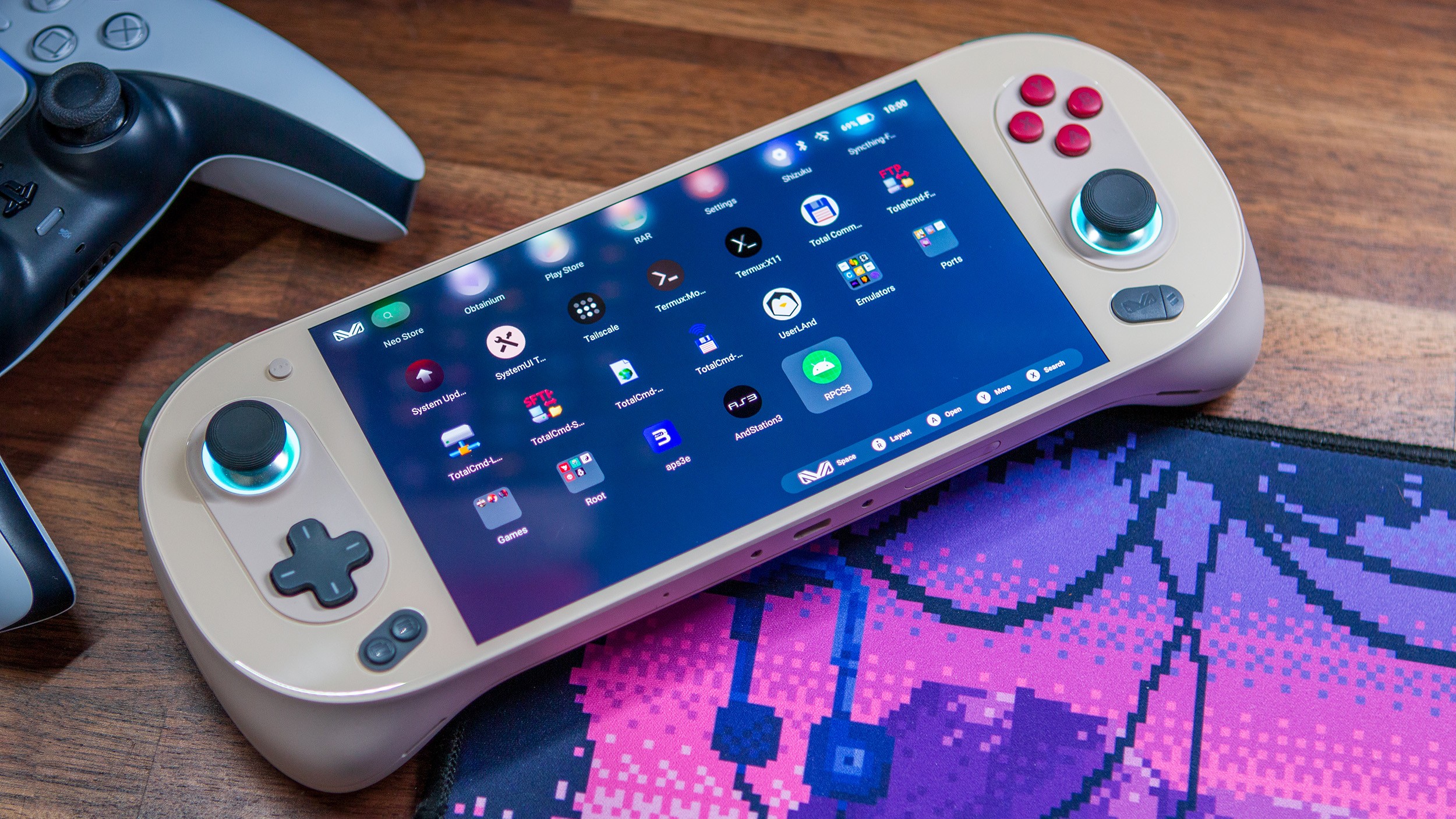 Handheld gaming device displays app icons on screen, resting on wooden surface beside PS5 controller. Handheld gaming device displays app icons on screen, resting on wooden surface beside PS5 controller.