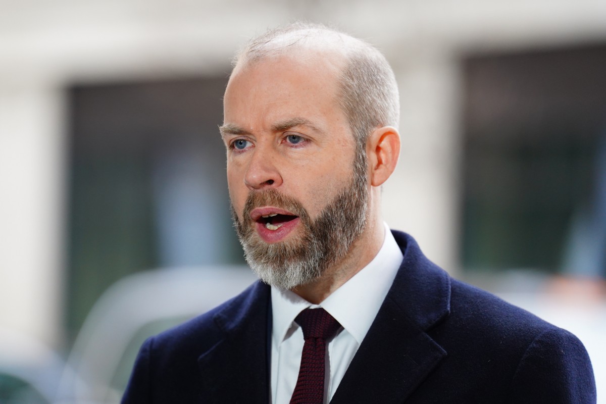 A bearded man in a dark suit and maroon tie speaks outdoors near blurred buildings. A bearded man in a dark suit and maroon tie speaks outdoors near blurred buildings.