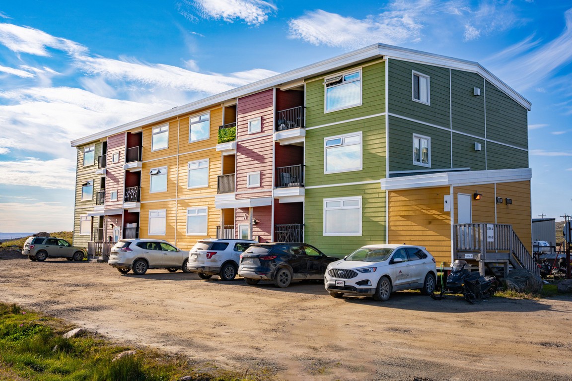 Colorful three-story apartment building with parked SUVs on a dirt lot under blue sky. Colorful three-story apartment building with parked SUVs on a dirt lot under blue sky.