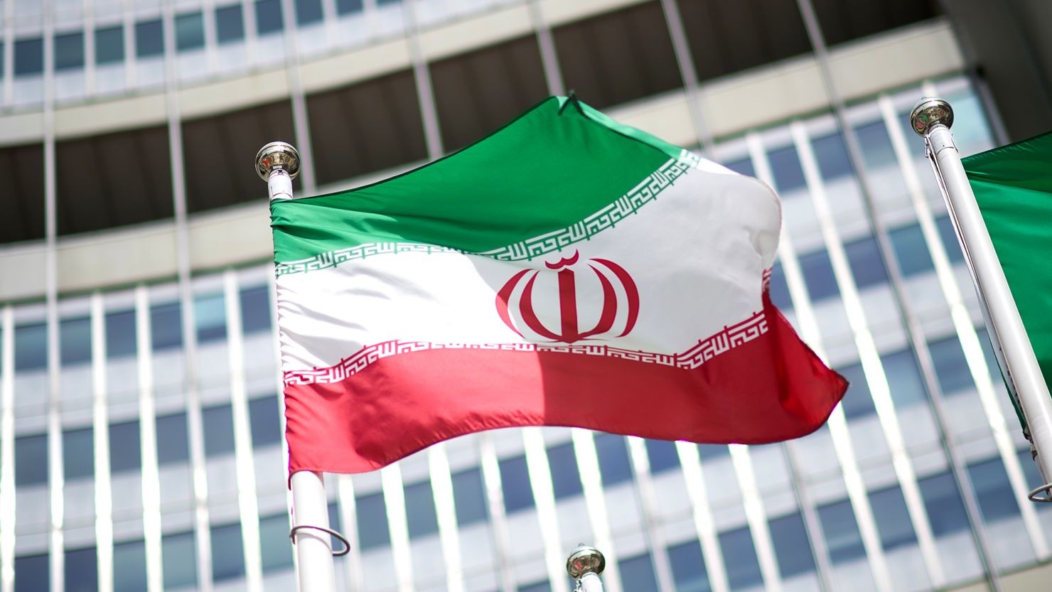 Iranian flag waves in front of a modern building with grid-patterned windows. Iranian flag waves in front of a modern building with grid-patterned windows.
