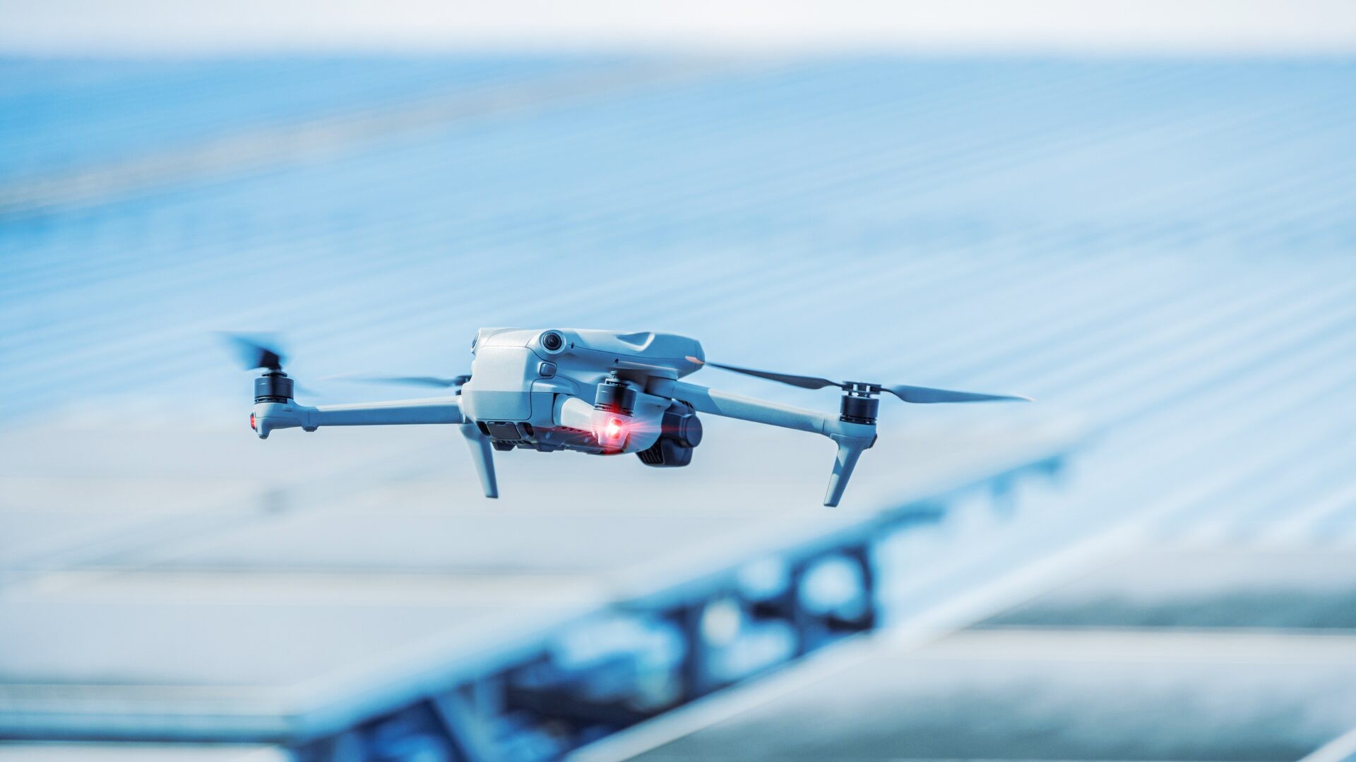 A white drone flies against a blue sky with blurred structures below. A white drone flies against a blue sky with blurred structures below.
