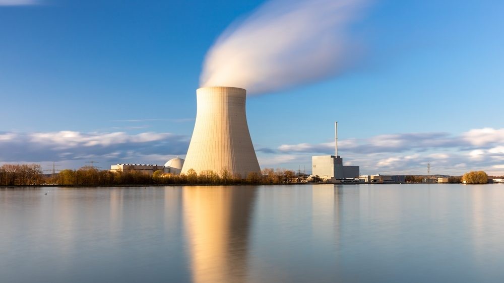 Nuclear power plant cooling tower emits steam over calm water under clear blue sky.