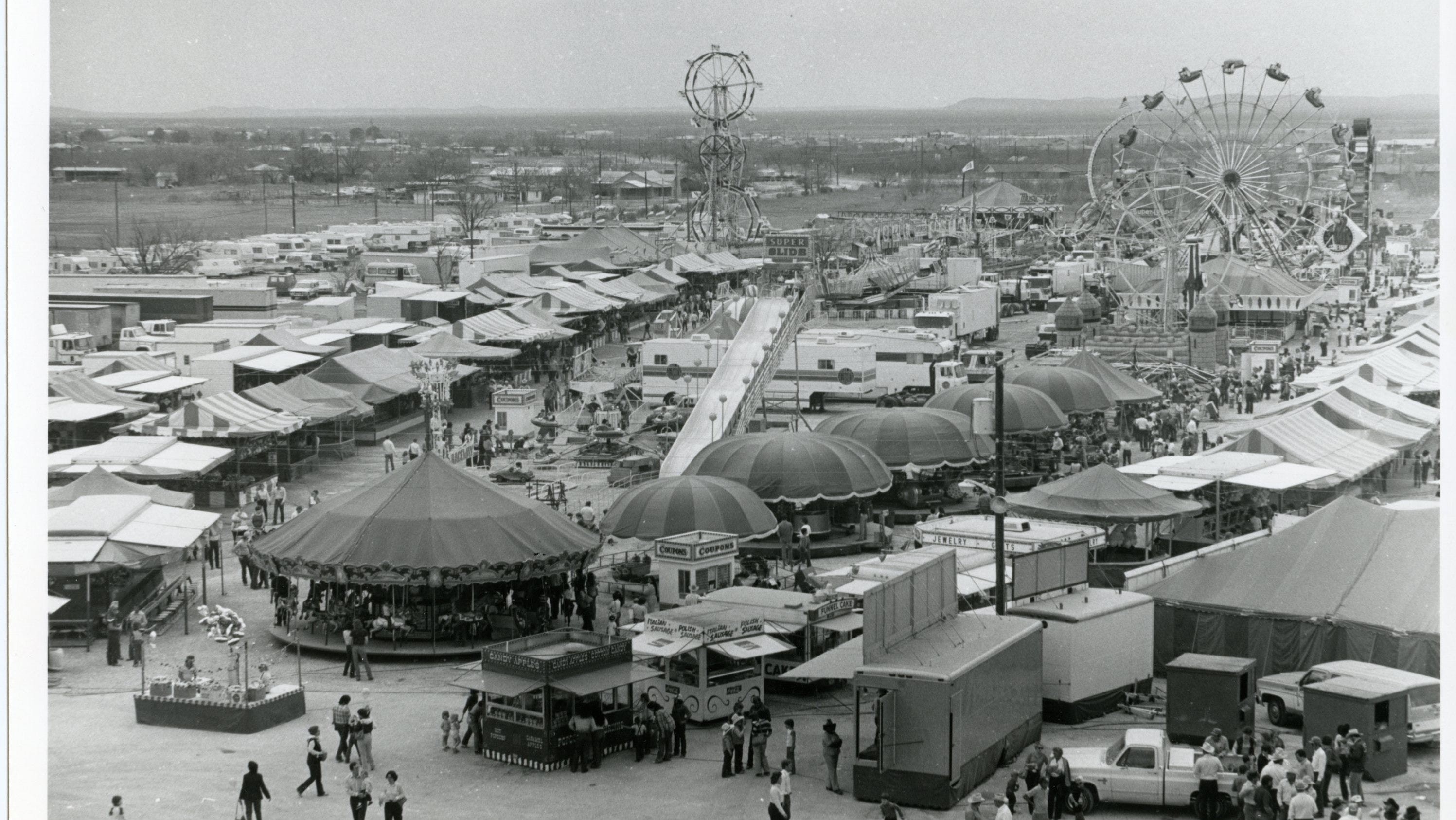 A large Ferris wheel stands at a busy fairground with people walking nearby on a sunny day.