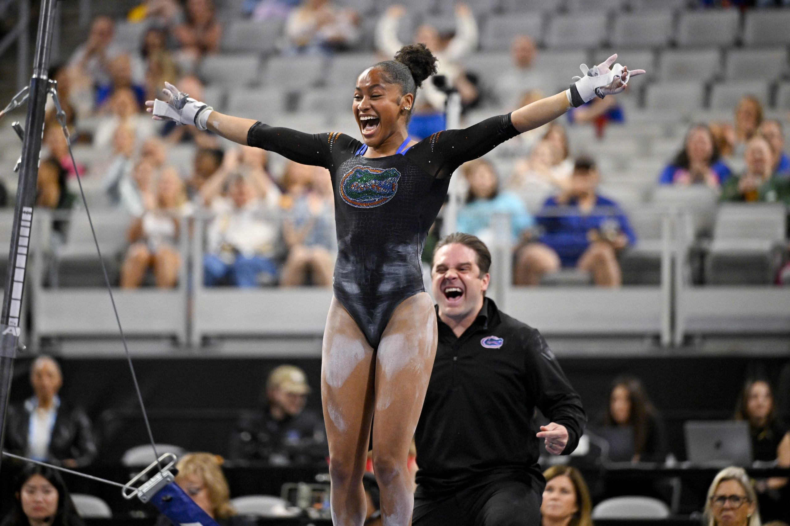 A female gymnast celebrates on balance beam in an indoor arena with spectators watching.
