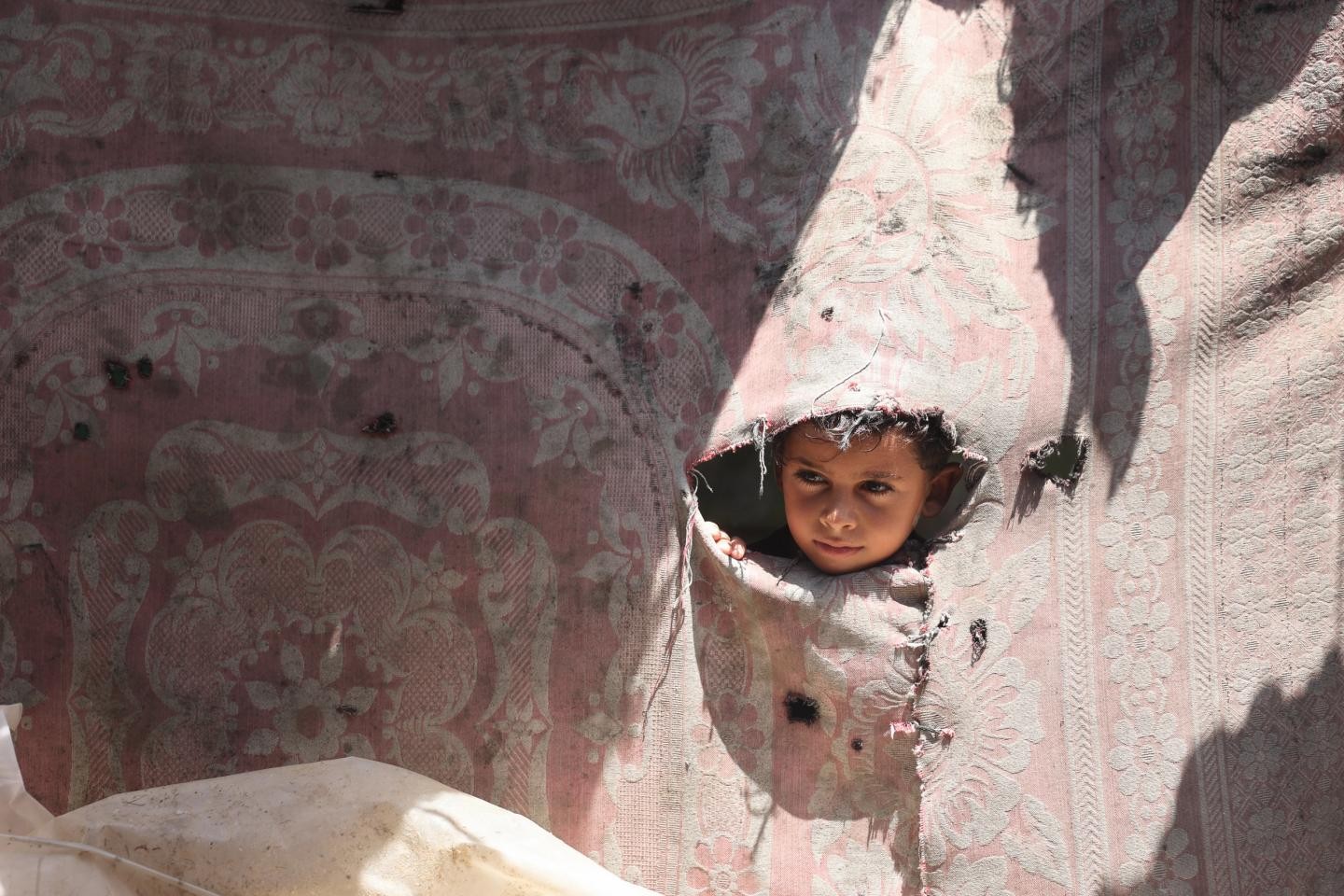 A child peers through a torn hole in a pink floral-patterned fabric wall. A child peers through a torn hole in a pink floral-patterned fabric wall.
