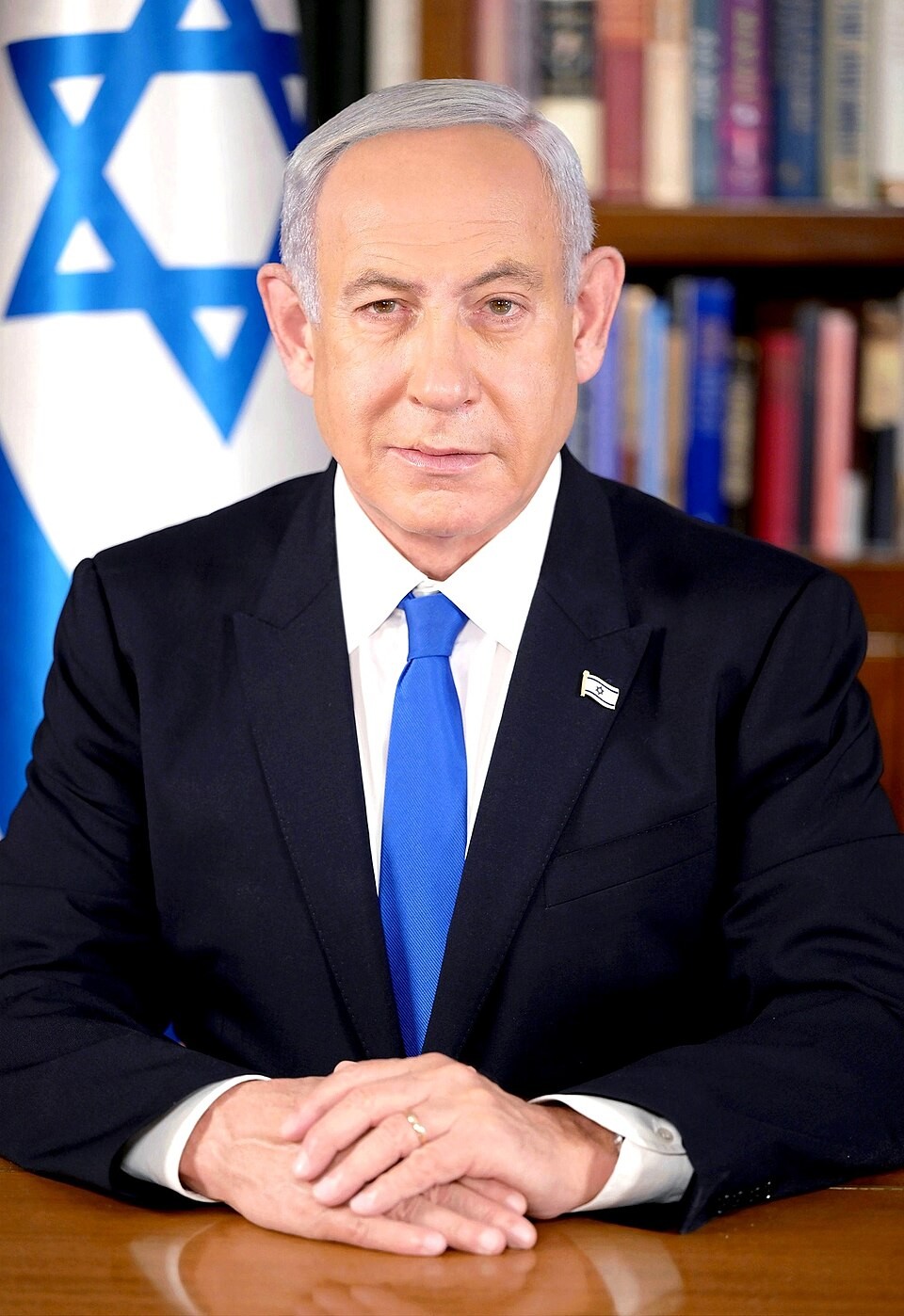 A man in a dark suit sits at a desk with hands clasped before an Israeli flag and bookshelf. A man in a dark suit sits at a desk with hands clasped before an Israeli flag and bookshelf.