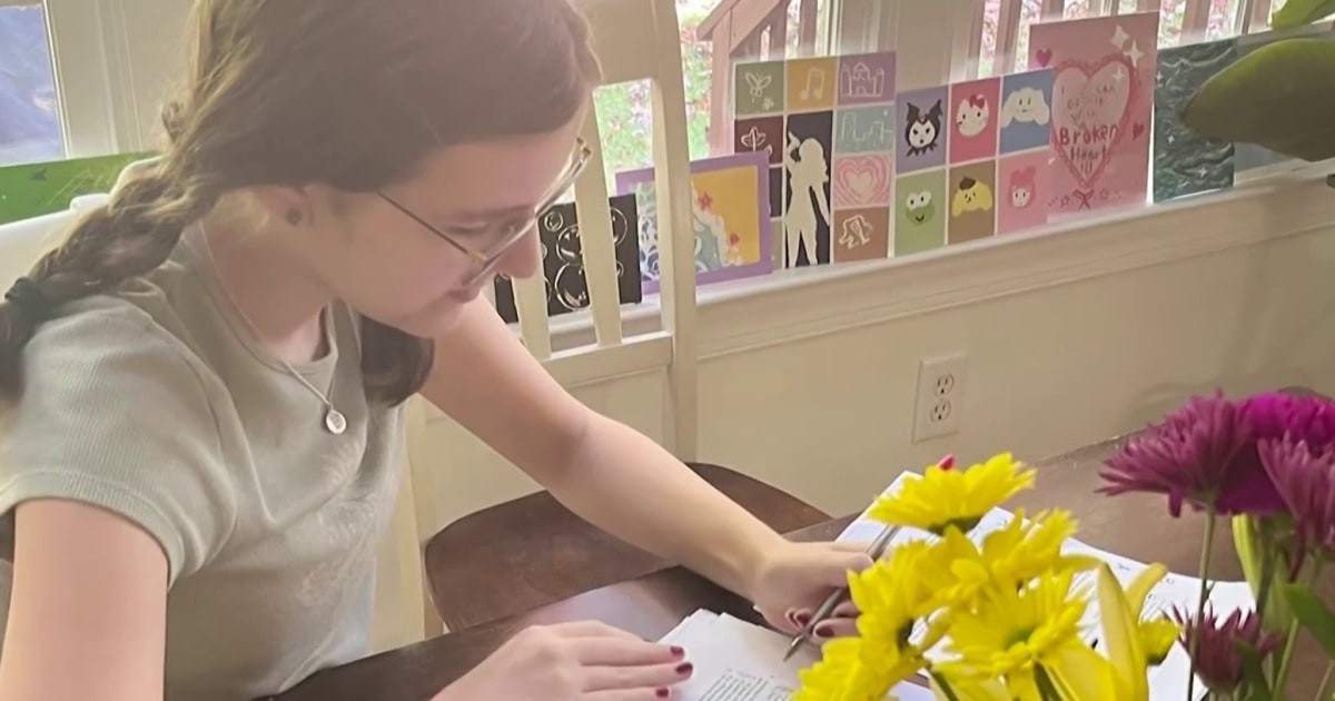 A woman with braids writes at a table near a window with colorful cards and flowers. A woman with braids writes at a table near a window with colorful cards and flowers.