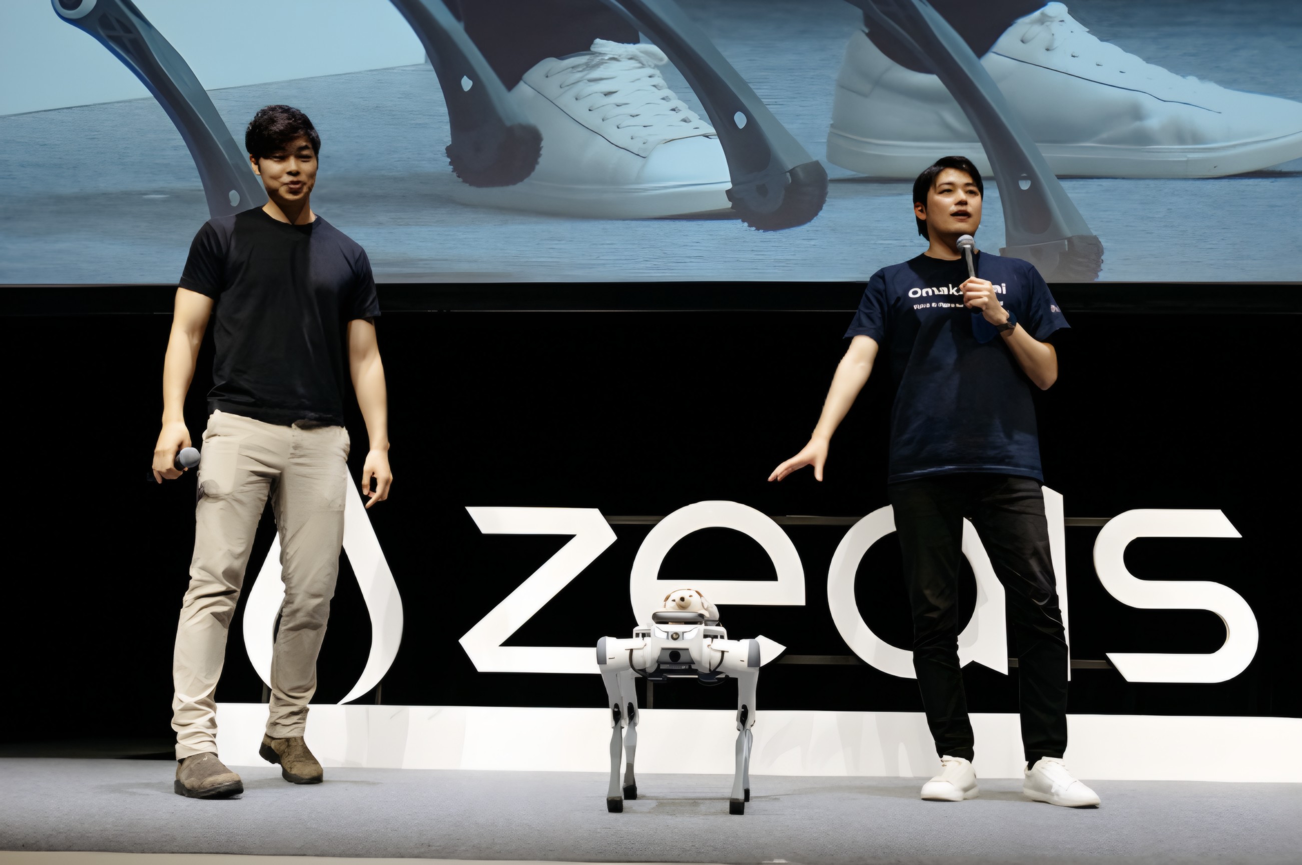 Two men stand on a stage with a quadruped robot, speaking into microphones before a "zeals" backdrop.