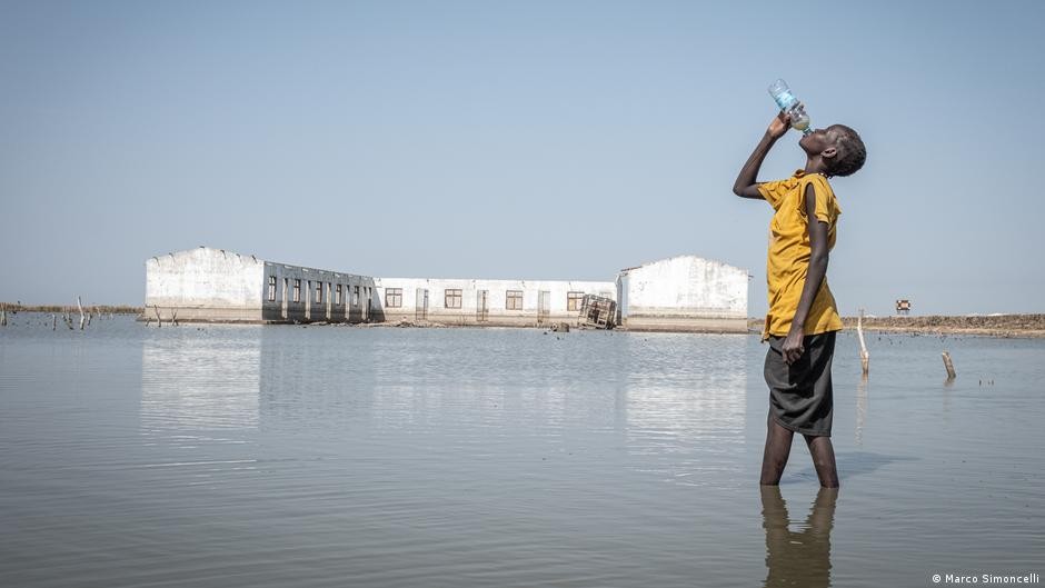A boy drinks from a bottle standing in shallow floodwater near submerged buildings. A boy drinks from a bottle standing in shallow floodwater near submerged buildings.