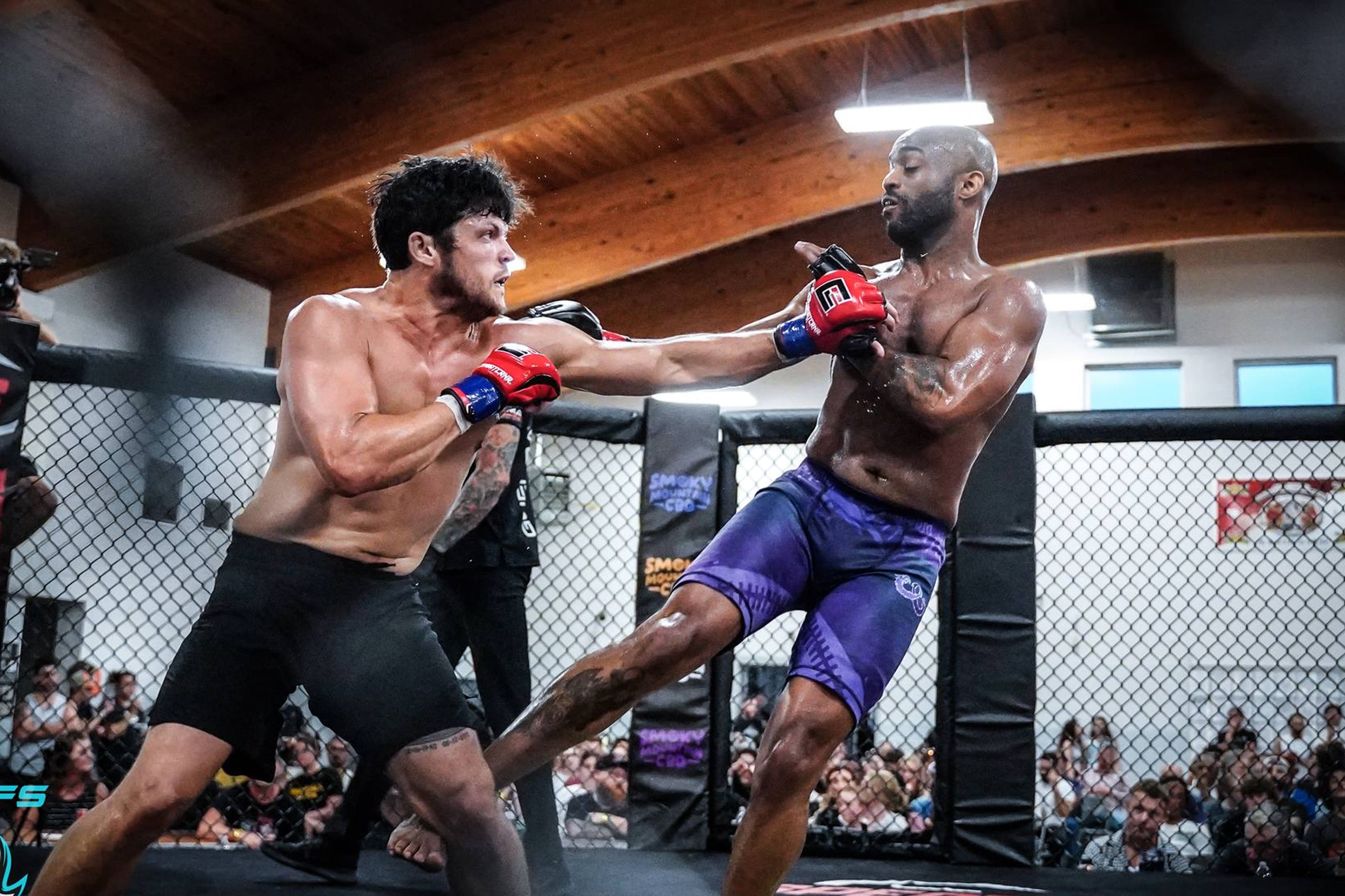 Two shirtless men fight inside an MMA cage with spectators watching.