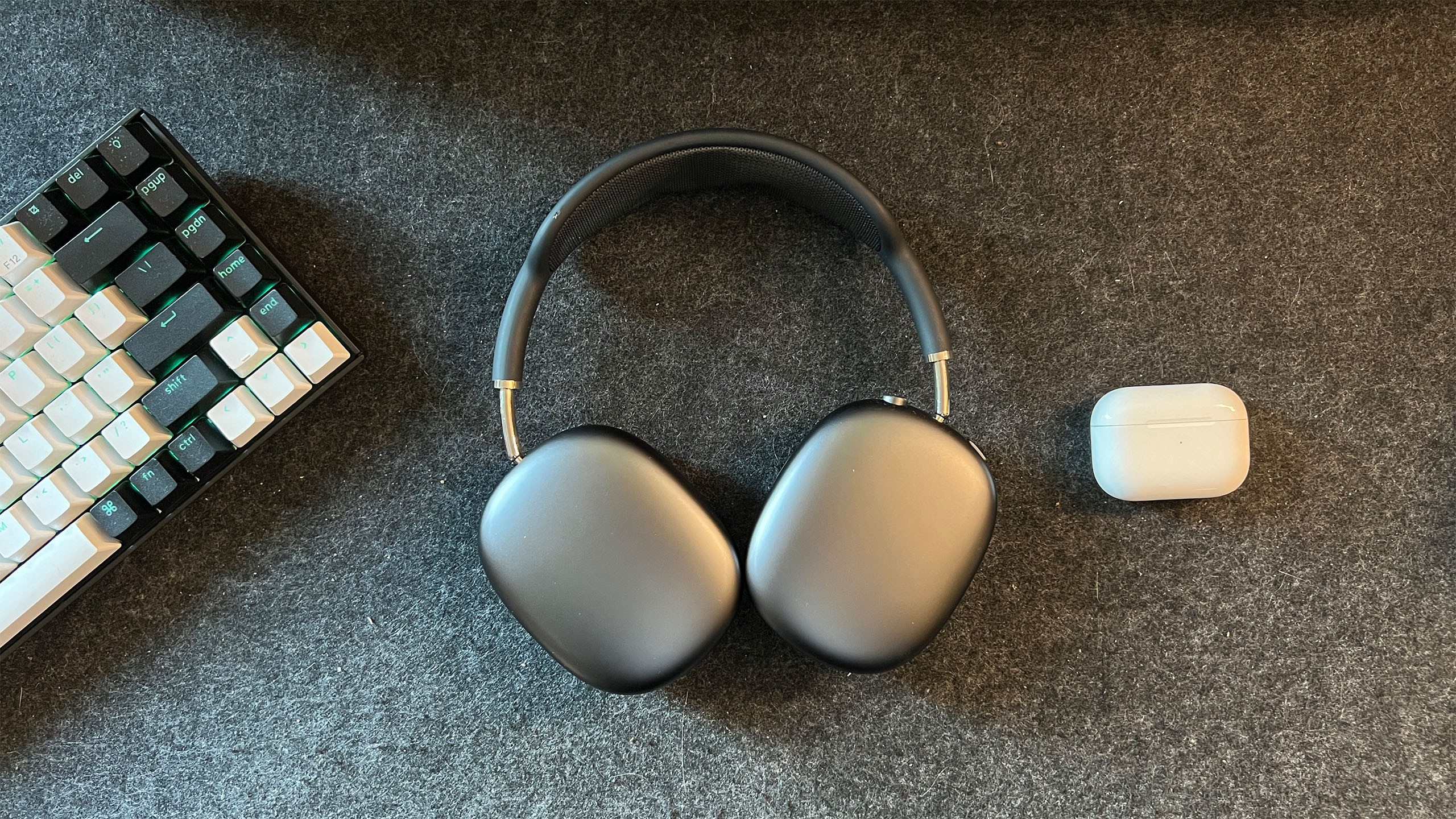 A mechanical keyboard, over-ear headphones, and white earbud case rest on a dark textured surface. A mechanical keyboard, over-ear headphones, and white earbud case rest on a dark textured surface.