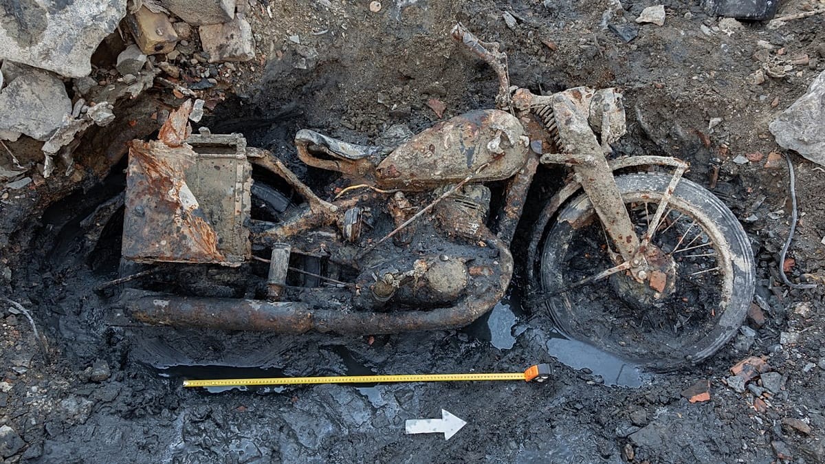 Rusted motorcycle lies buried in muddy debris with a yellow measuring tape visible. Rusted motorcycle lies buried in muddy debris with a yellow measuring tape visible.