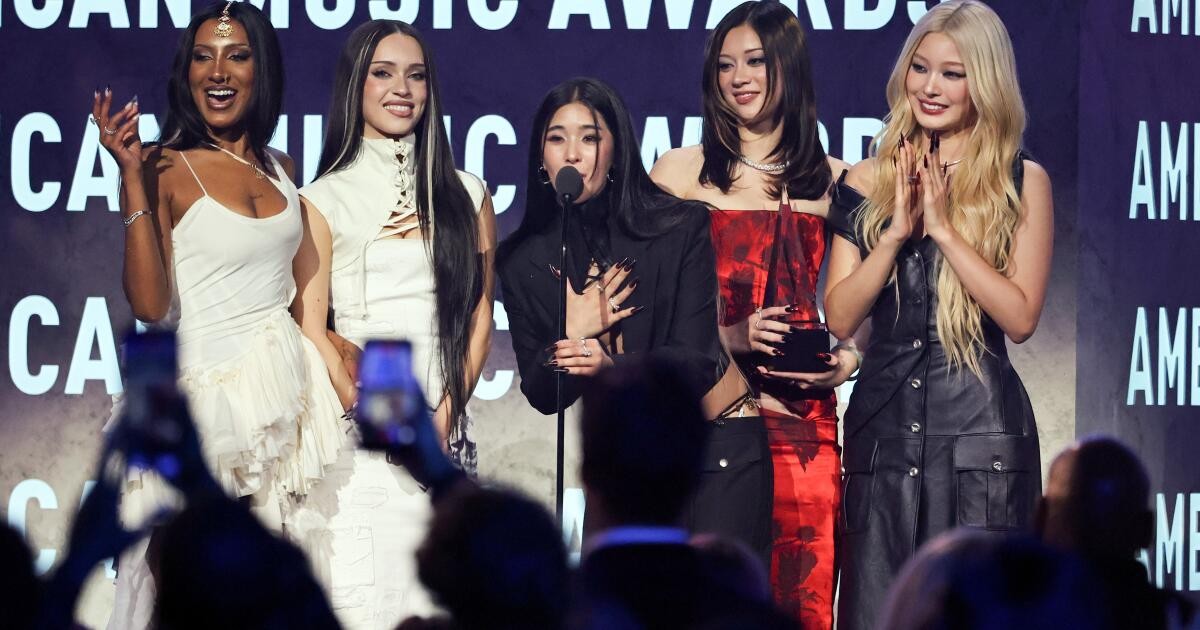 Five women pose on American Music Awards stage; one speaks into microphone while holding award. Five women pose on American Music Awards stage; one speaks into microphone while holding award.