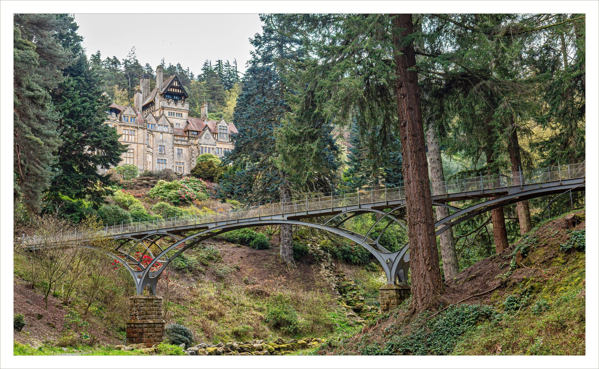 An arched pedestrian bridge spans a forested hillside near a large stone mansion. An arched pedestrian bridge spans a forested hillside near a large stone mansion.