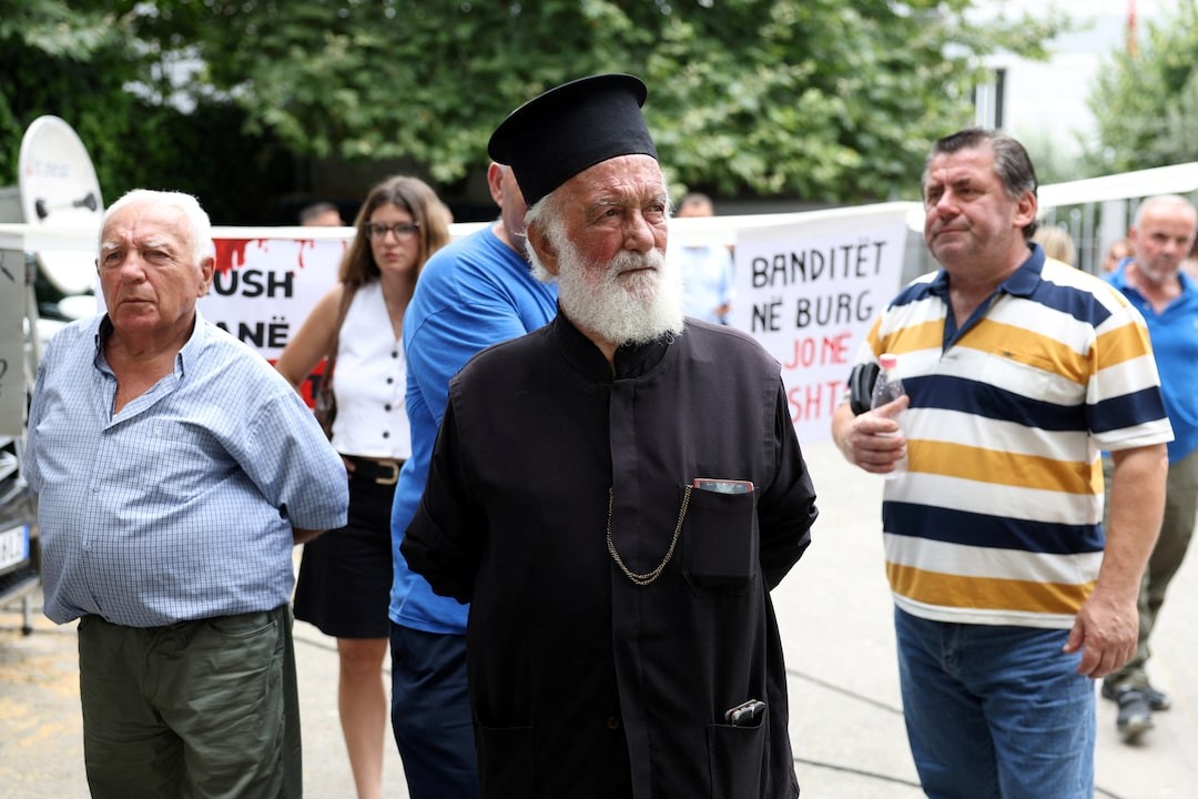 Elderly man with white beard and black hat stands at an outdoor protest with Albanian-language banners.