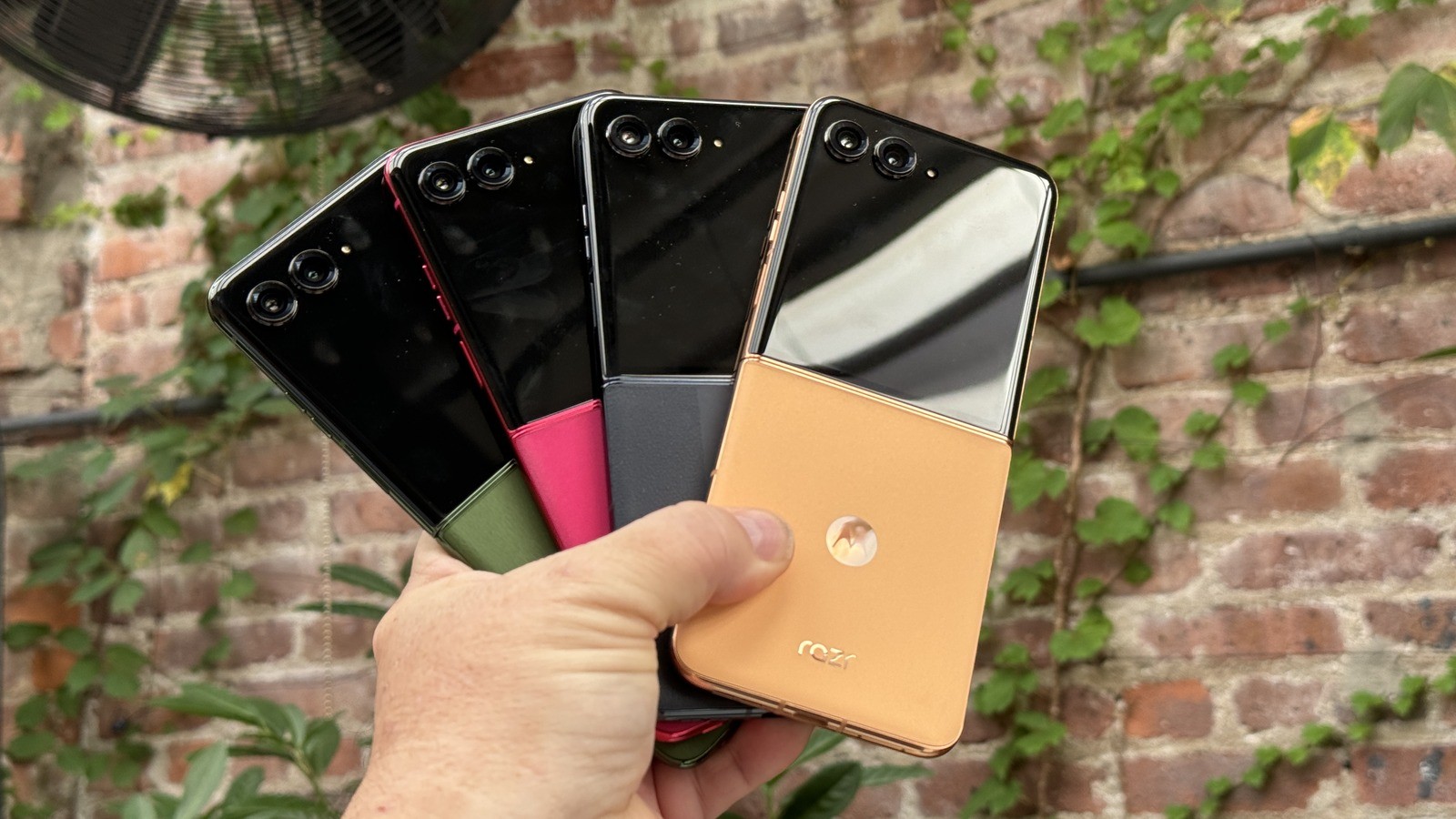 A hand holds several Motorola Razr flip phones against a brick wall with ivy.