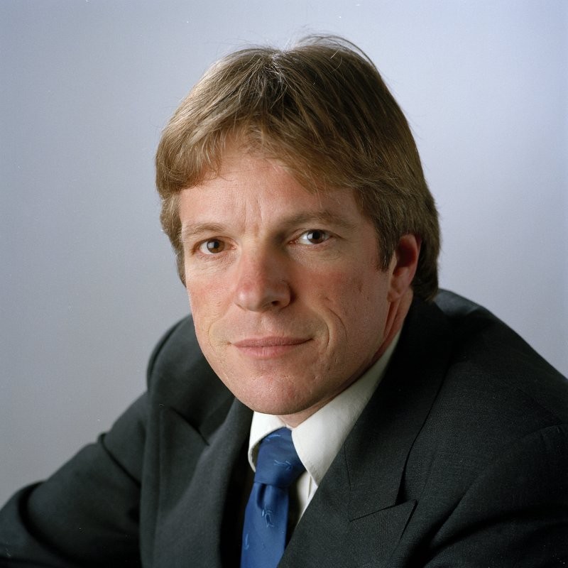 A man in a dark suit and blue tie poses against a plain light gray backdrop. A man in a dark suit and blue tie poses against a plain light gray backdrop.