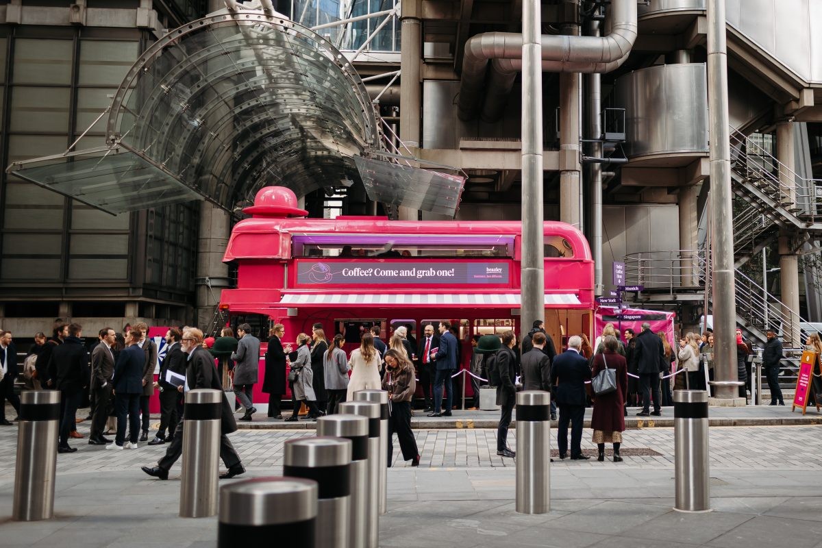 Red double-decker coffee kiosk in urban plaza; crowd gathers around it. Red double-decker coffee kiosk in urban plaza; crowd gathers around it.