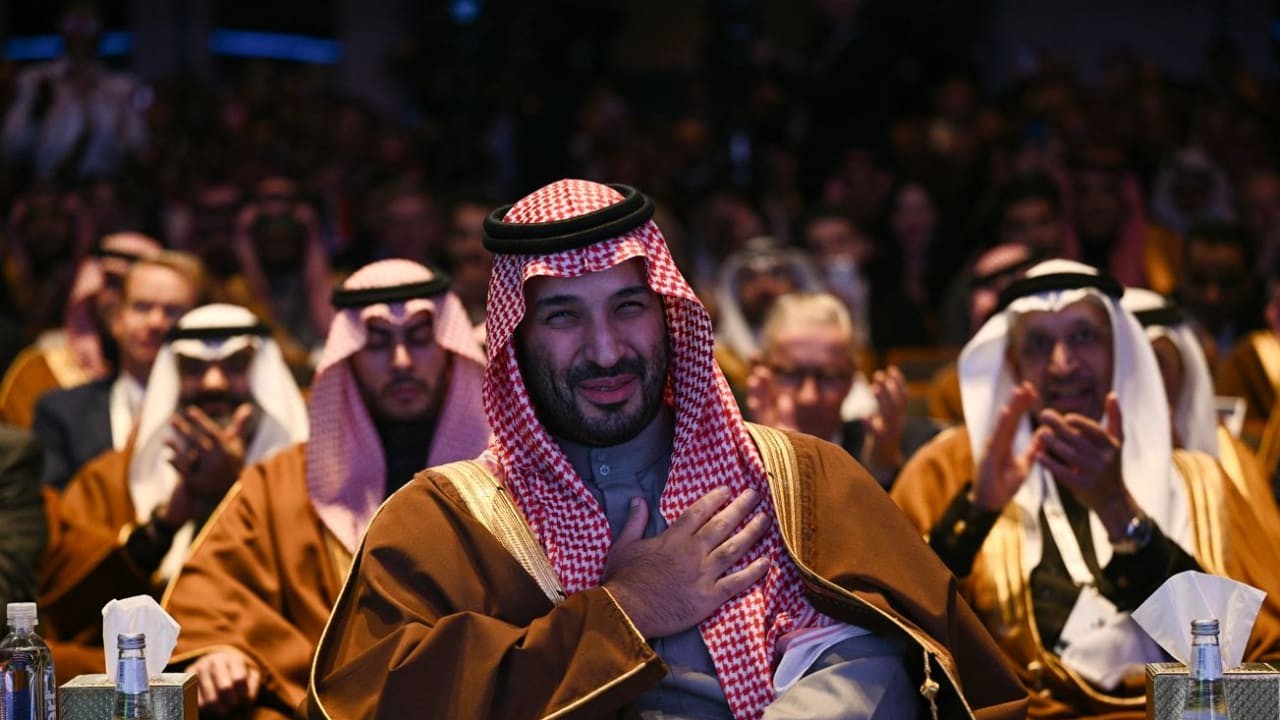 Men in traditional Arabian attire clap at a formal indoor event.