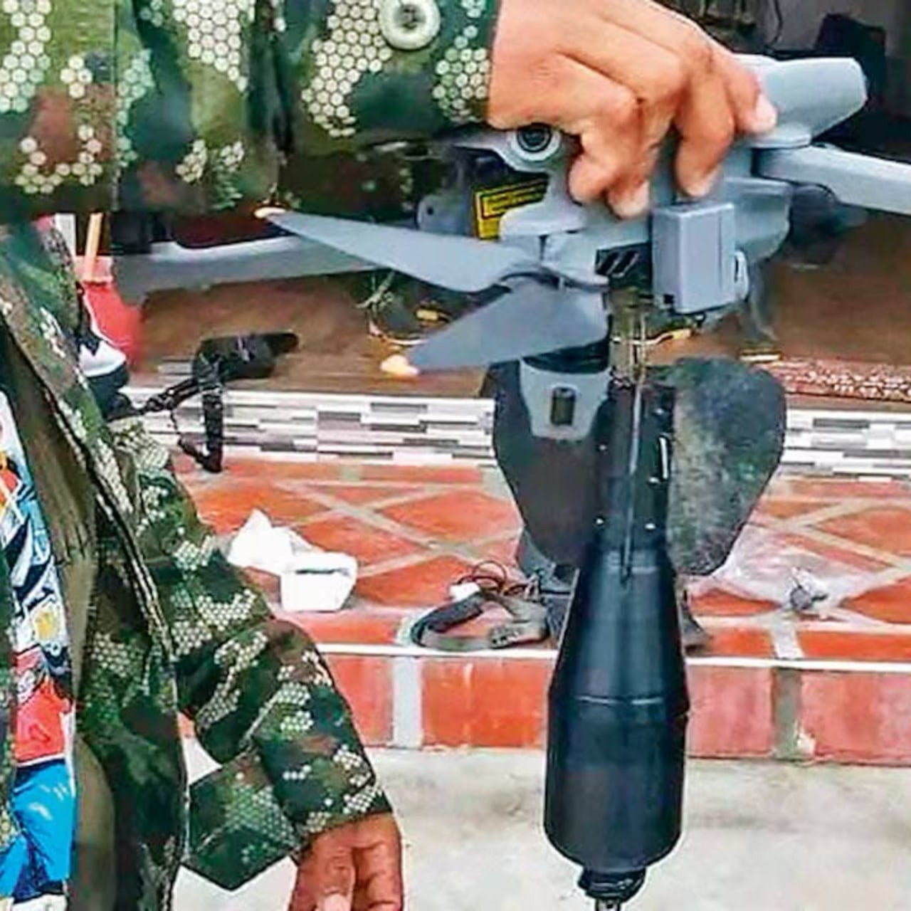 Person in military camouflage holds drone with attached black payload indoors on tiled floor. Person in military camouflage holds drone with attached black payload indoors on tiled floor.