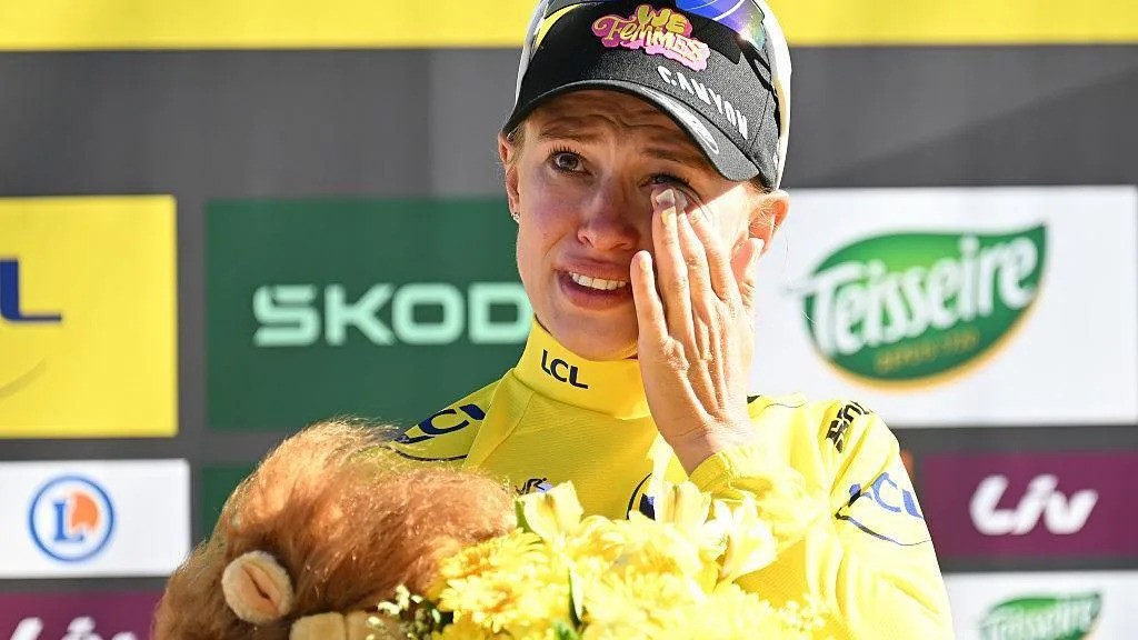 Cyclist in yellow LCL jersey holds flowers and teddy bear, wiping tear at sponsor-adorned podium.
