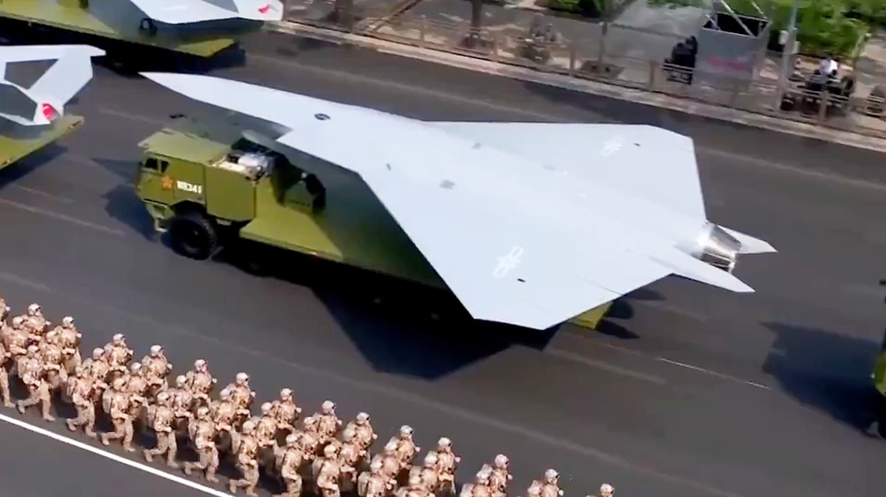 A large white aircraft is transported by a green military truck while soldiers march on a street. A large white aircraft is transported by a green military truck while soldiers march on a street.