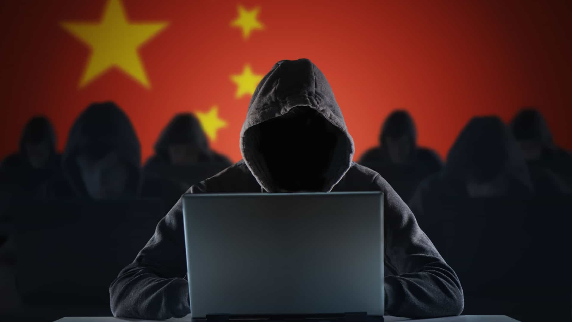 Hooded figure uses laptop before Chinese national flag backdrop. Hooded figure uses laptop before Chinese national flag backdrop.