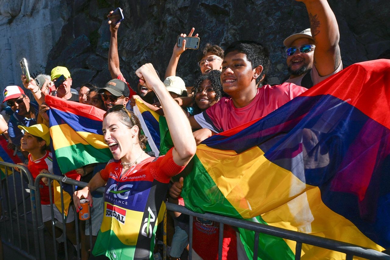 Spectators wave rainbow flags and cheer at an outdoor event near a rocky wall. Spectators wave rainbow flags and cheer at an outdoor event near a rocky wall.