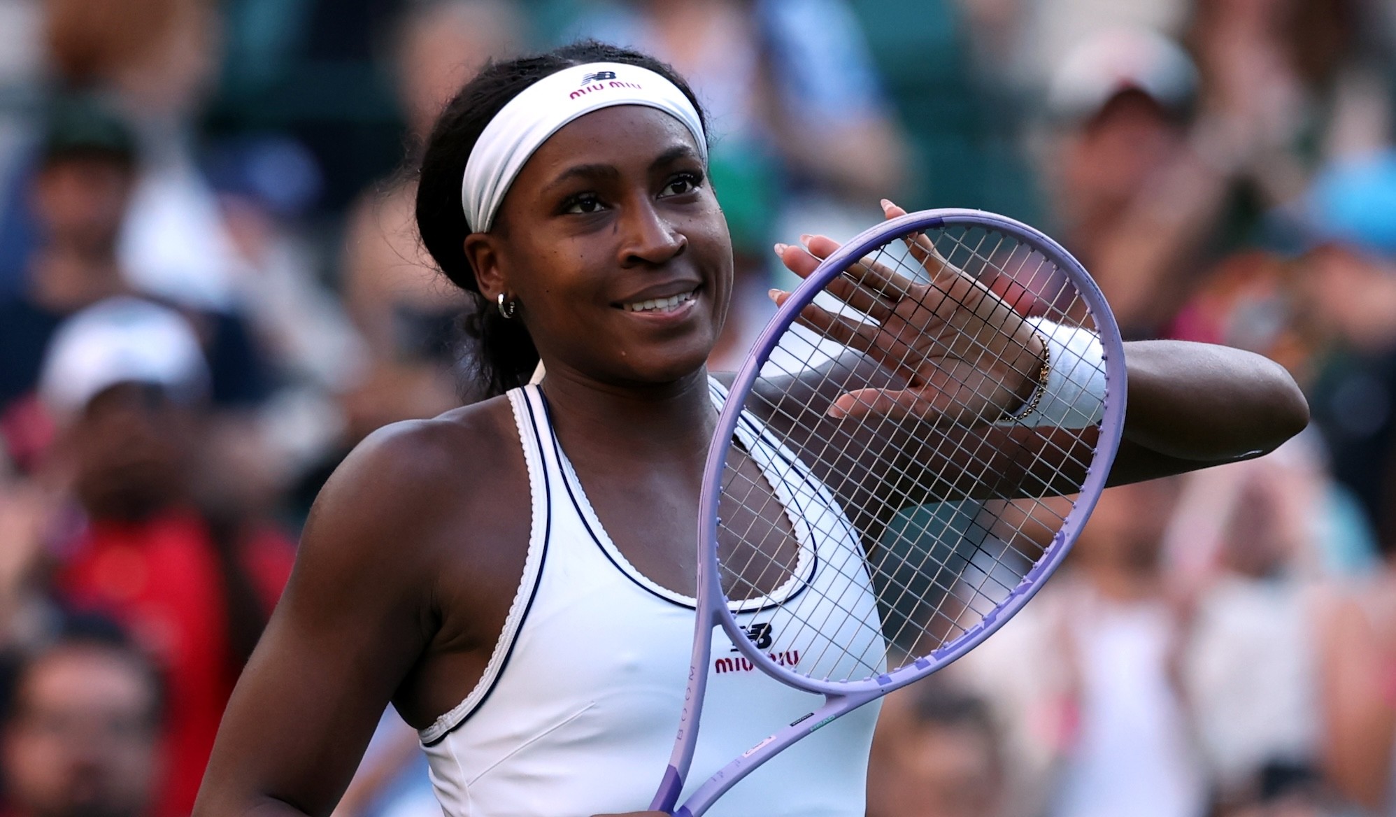 A female tennis player holds a purple racket and waves to spectators in a stadium. A female tennis player holds a purple racket and waves to spectators in a stadium.