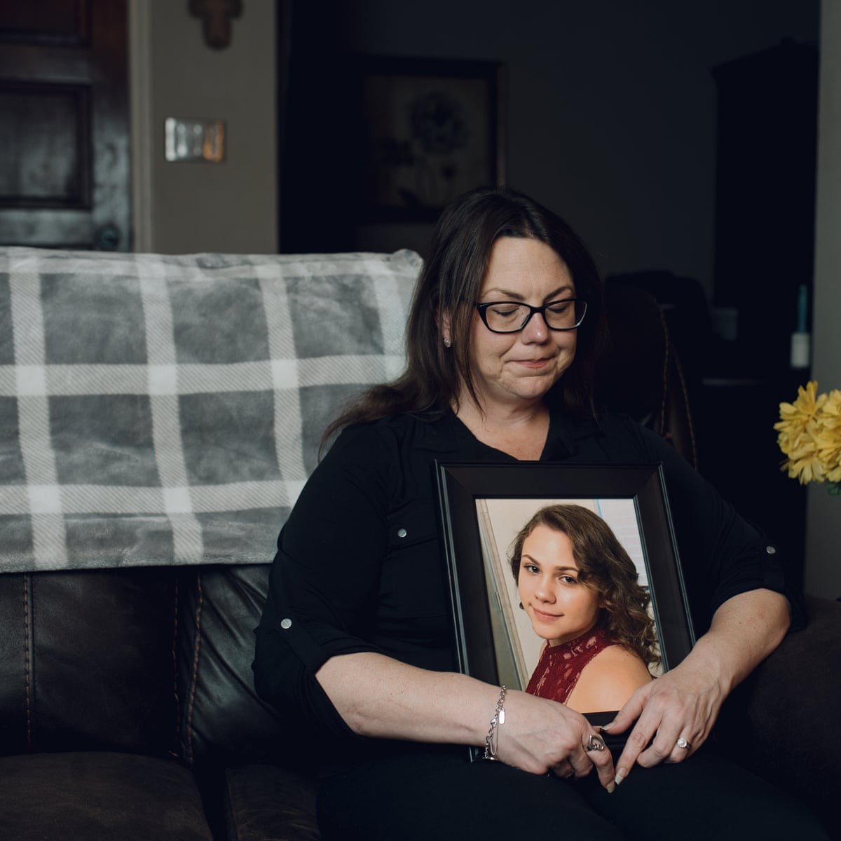 Woman holds framed photograph on dark leather sofa in dimly lit living room.