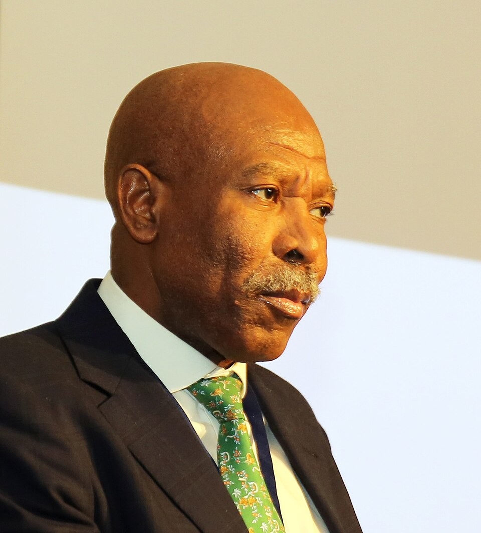 A bald man in a dark suit and green patterned tie looks sideways indoors.