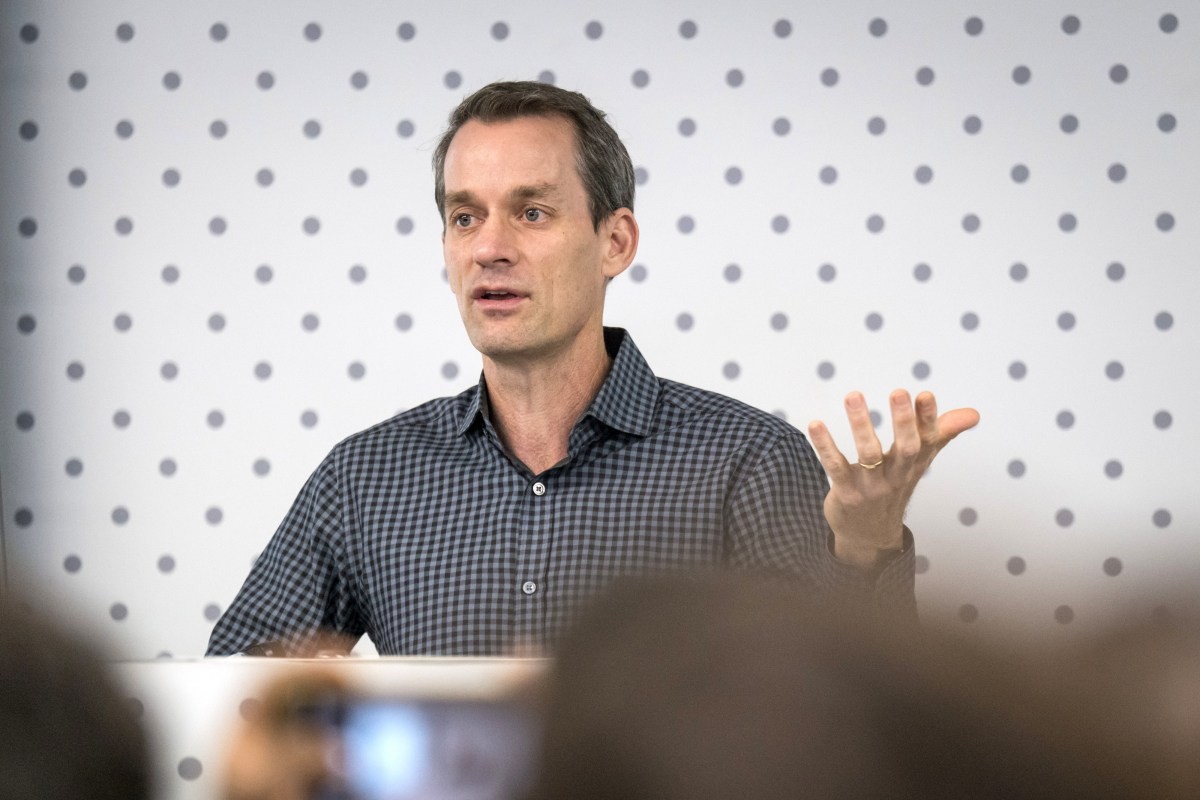 A man in a checkered shirt speaks and gestures with his right hand against a polka-dot wall. A man in a checkered shirt speaks and gestures with his right hand against a polka-dot wall.