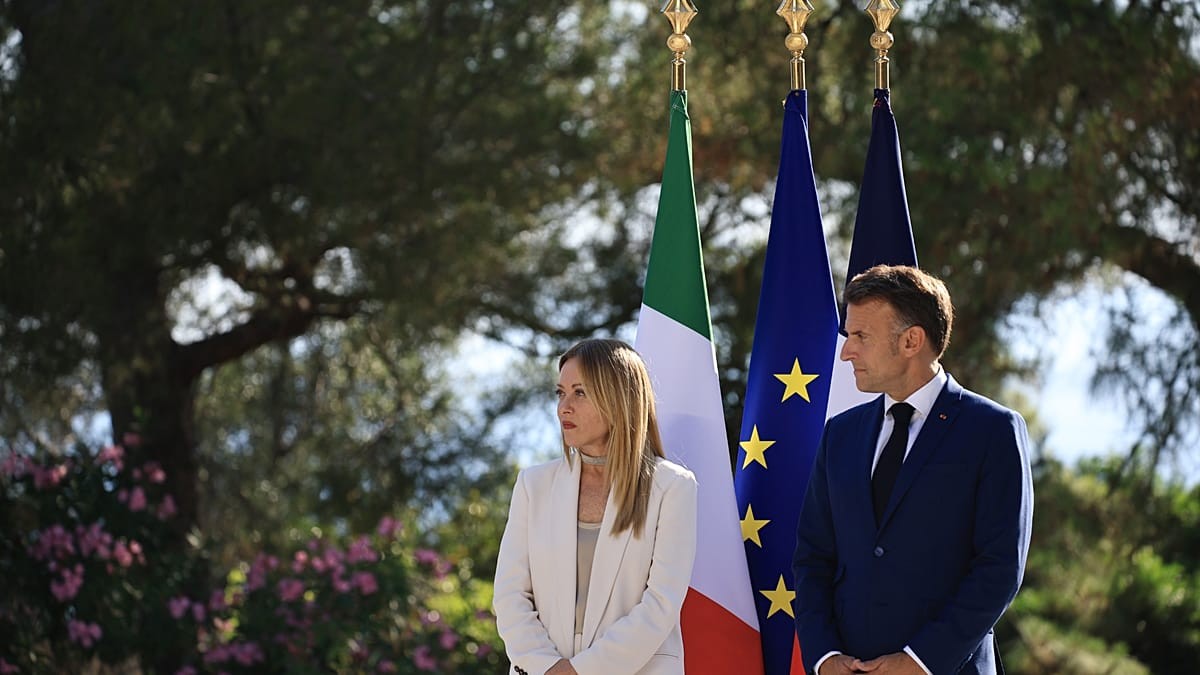 A woman in white suit and a man in blue suit stand before three flags outdoors.