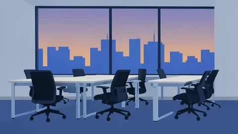 A row of empty office desks with chairs pushed in, set against a window overlooking a city skyline at dusk.