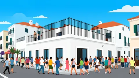 A crowded street corner in a Mediterranean coastal city with white buildings and a high perimeter fence in the background