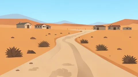 A dusty, unpaved road winding through a dry, arid landscape with scattered low-rise buildings in the distance