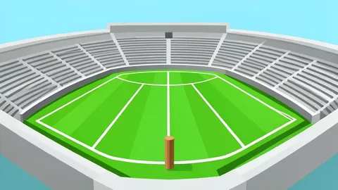 A lush green cricket pitch with white creases and stumps, surrounded by empty stadium stands under a clear sky.